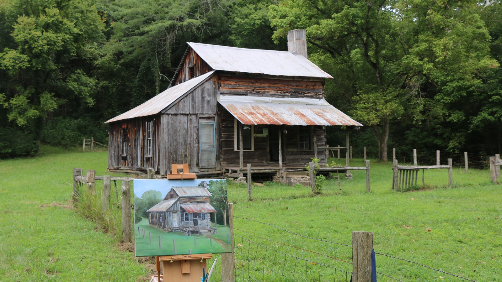 A plein air painting of a cabin sits on an easel in front of the cabin that inspired the painting.