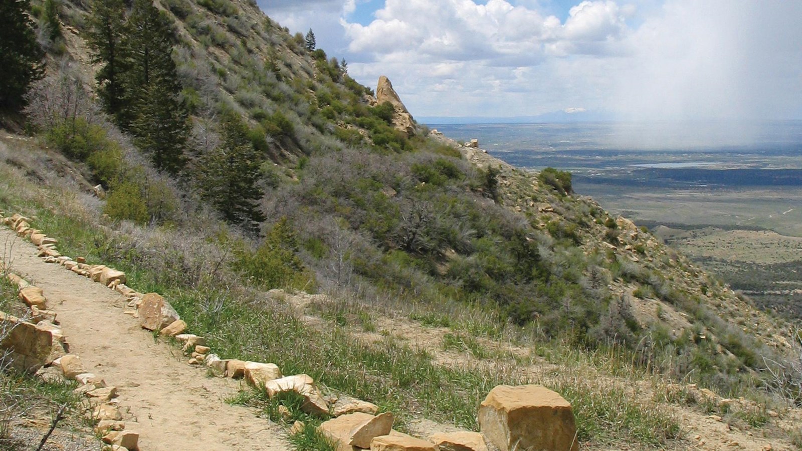 Dirt trail, lined with stones, traverses a ridge with view of a green valley in the distance below.