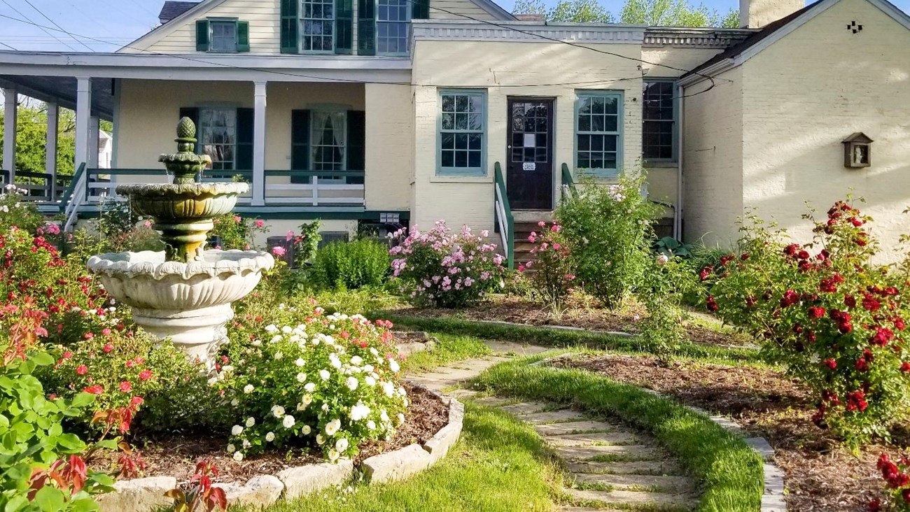 A rose garden in full bloom in front of a large, yellow home. A circular fountain sits in the middle