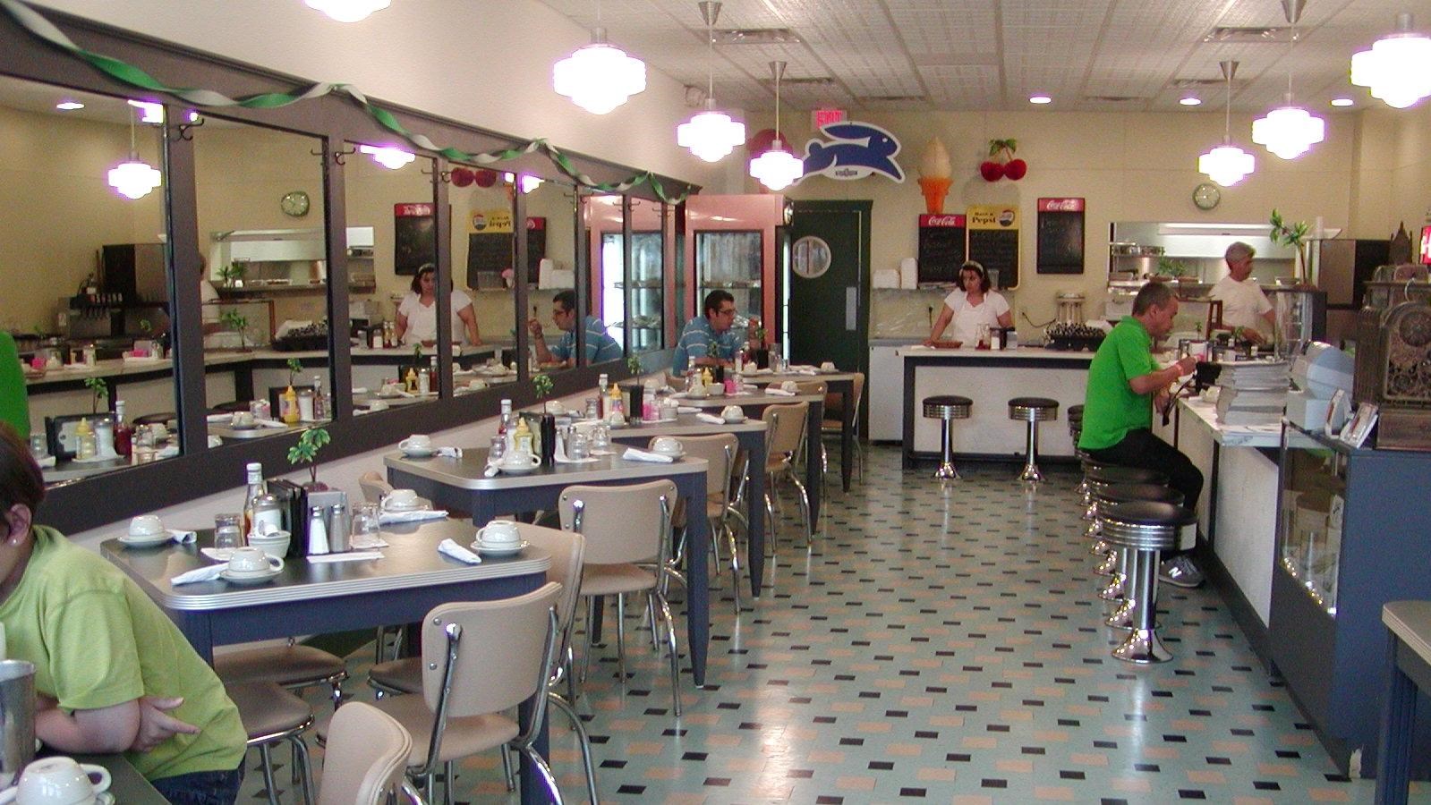 The inside of a cafe, with white and blue floors, a bar, and tables with people sitting at them.