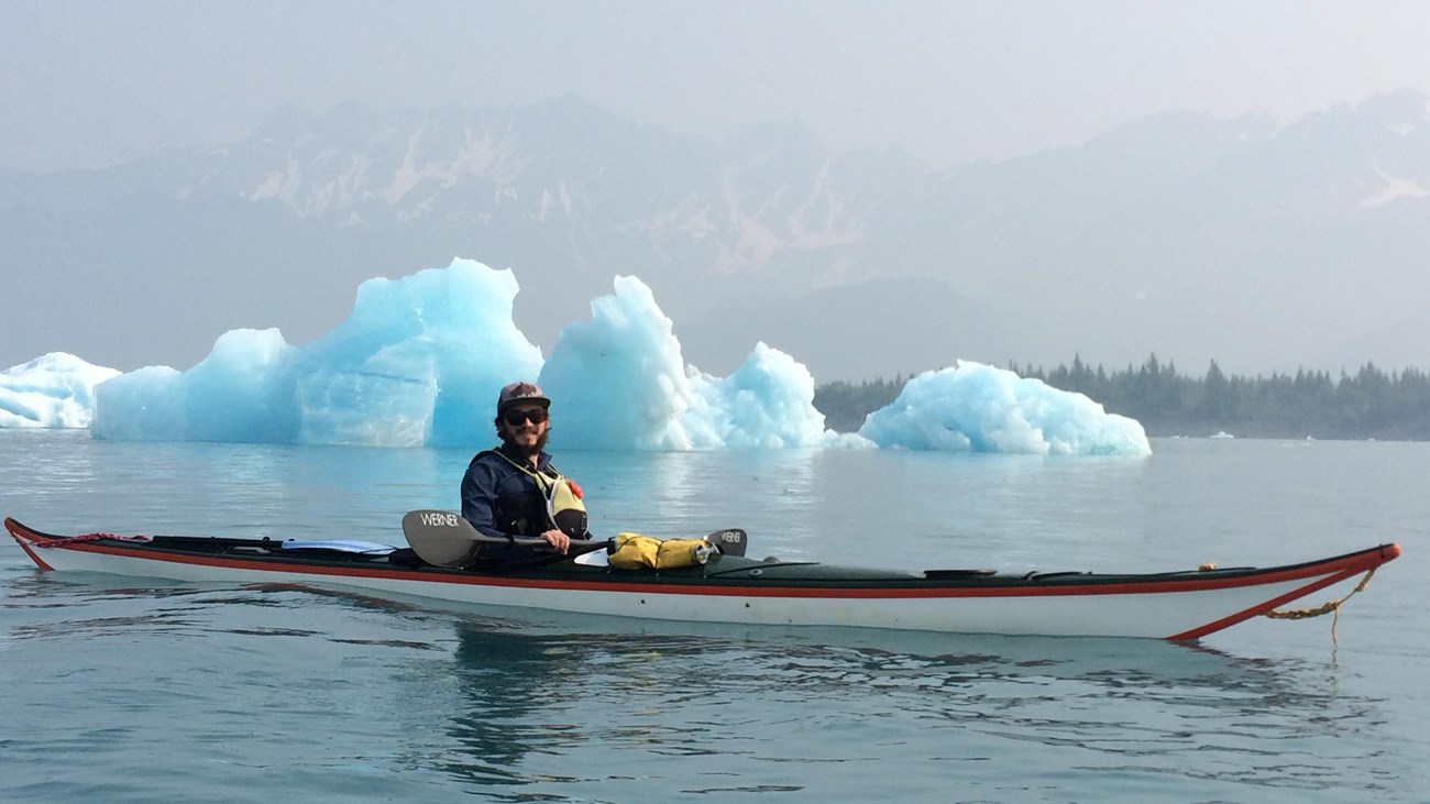 A kayaker is in front of a large blue colored iceberg in the background.