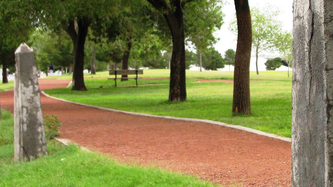 A red gravel trail that has trees and grass surrounding it with 2 boundary fence posts.