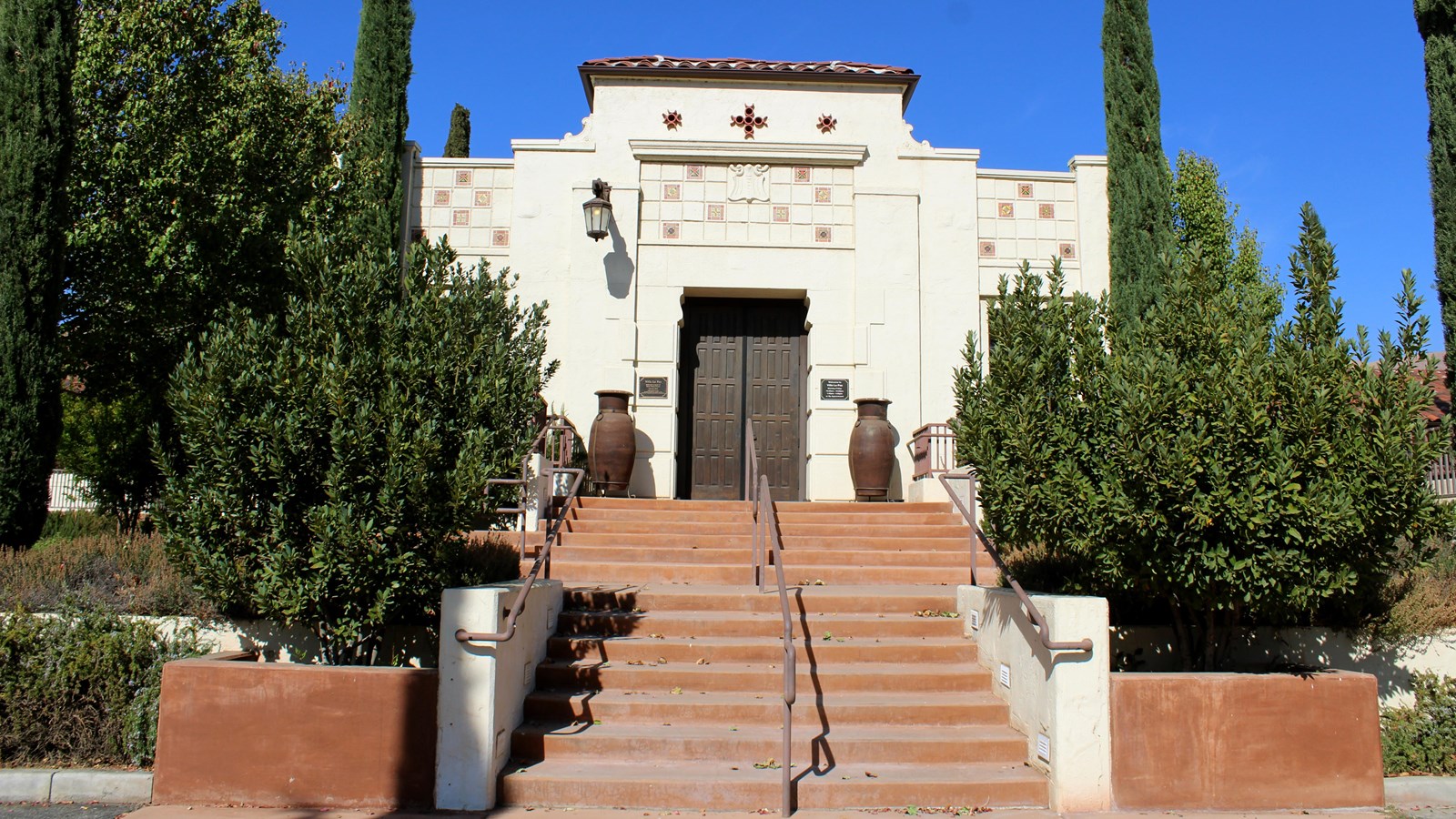 A white building with a red tile roof and stairs leading up to a black door. Trees line the stairs