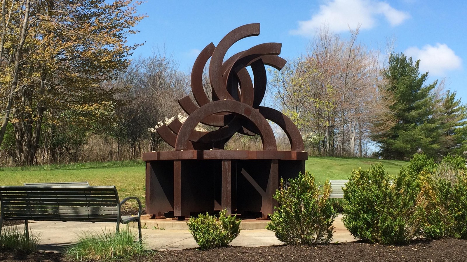 Rust-colored metal sculpture depicting broken Olympic rings surrounded by a circular cement walkway.
