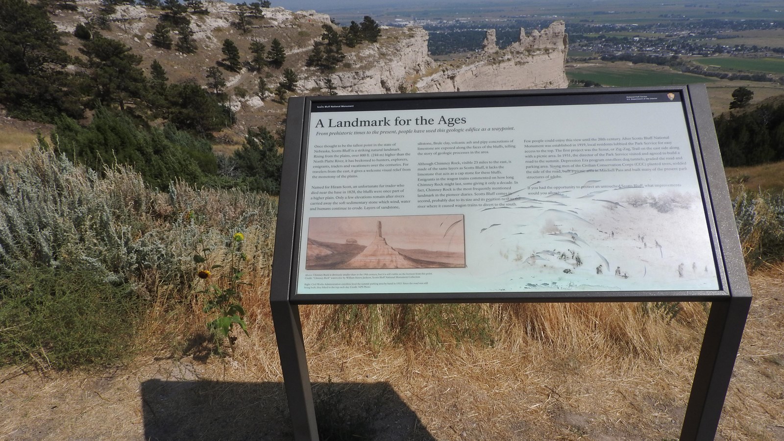 An interpretive wayside panel is seen with a portion of a sandstone bluff behind it.