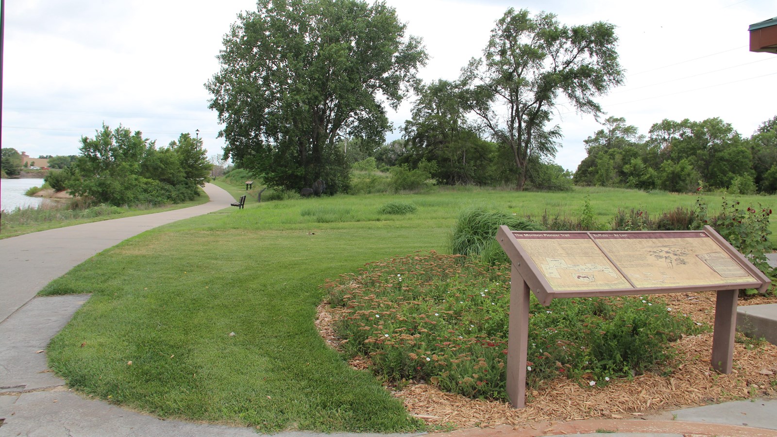Tan wayside panel shows Mormon Pioneer Trail. Grass, flowers, trees, and a concrete path surround. 