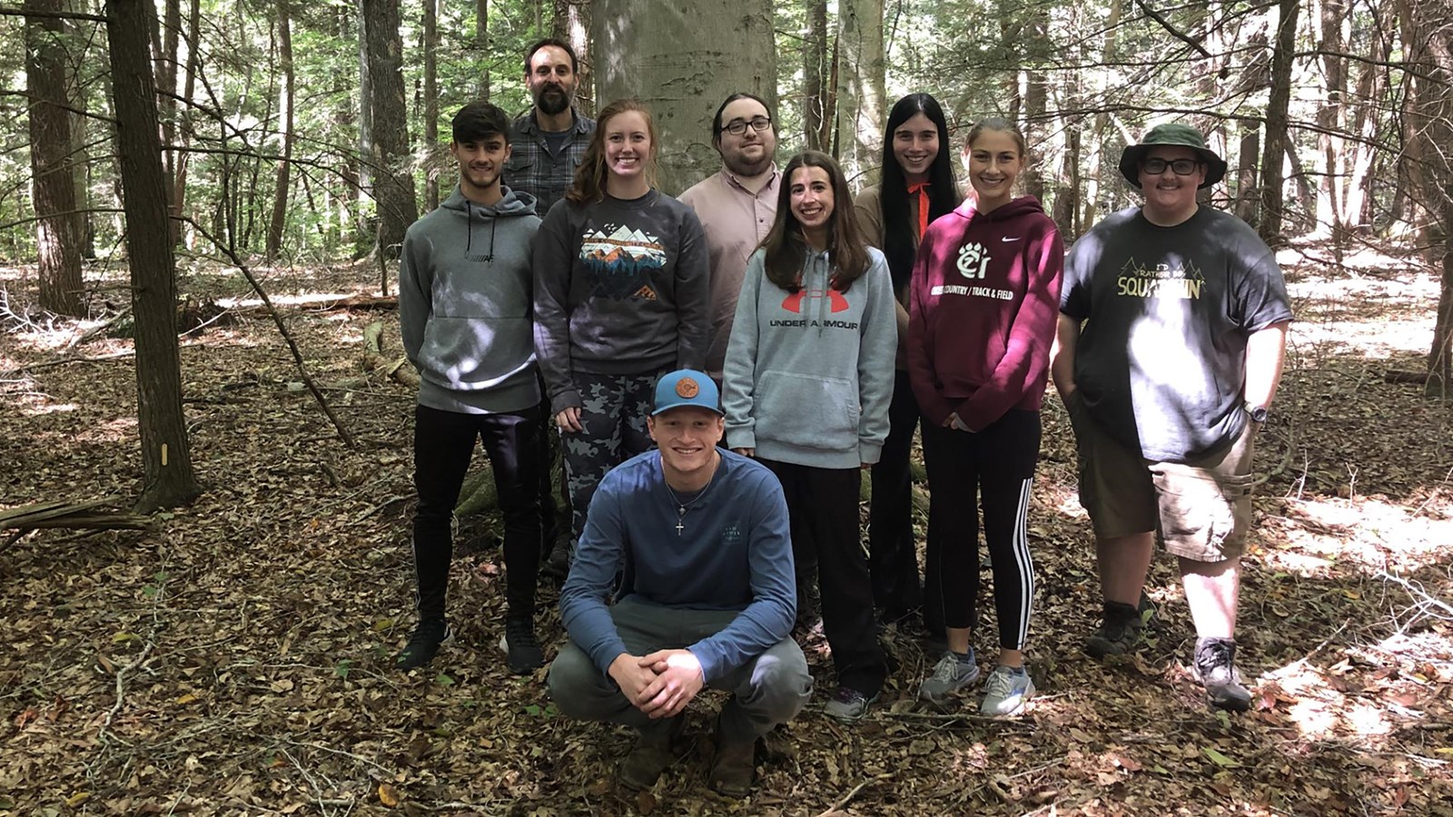 4 young women, 4 young men, and an older man posing and smiling in front of a large tree in a forest