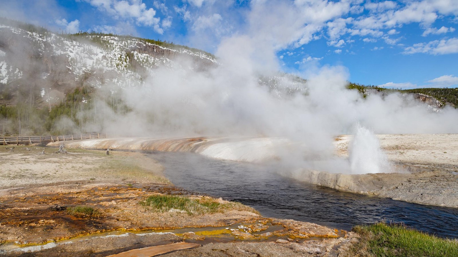 A small geyser erupts on the edge of a river flowing through a thermal basin.