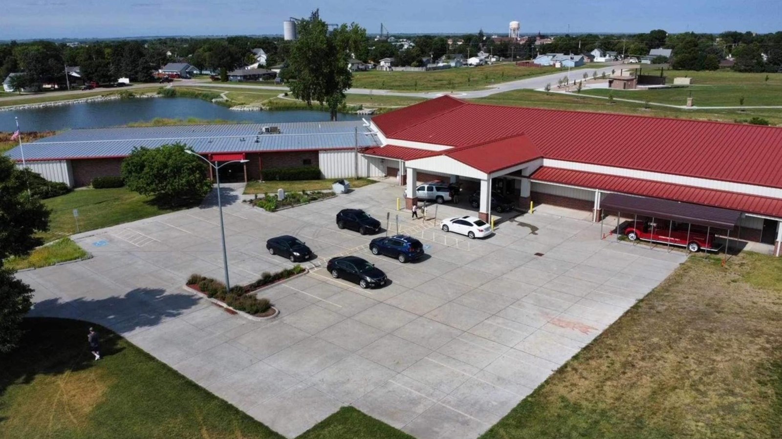 Aerial view of museum, parking lot with cars, red roof, fire truck, lake, trees, blue sky.