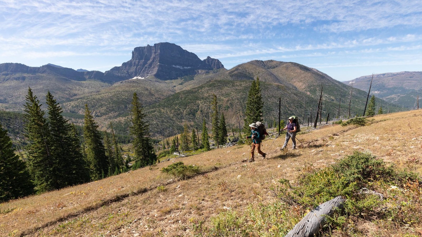 Two backpackers walk along a trail with a mountain peak in the background.