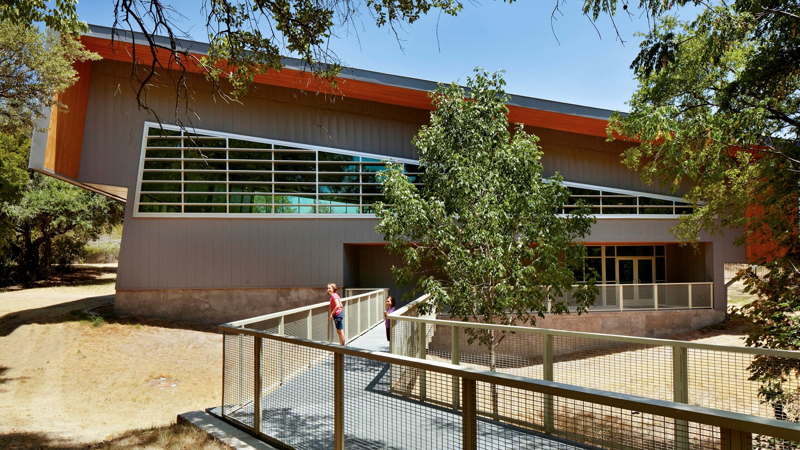 People stand on an elevated walkway leading to a large building with many windows and slanted roof.