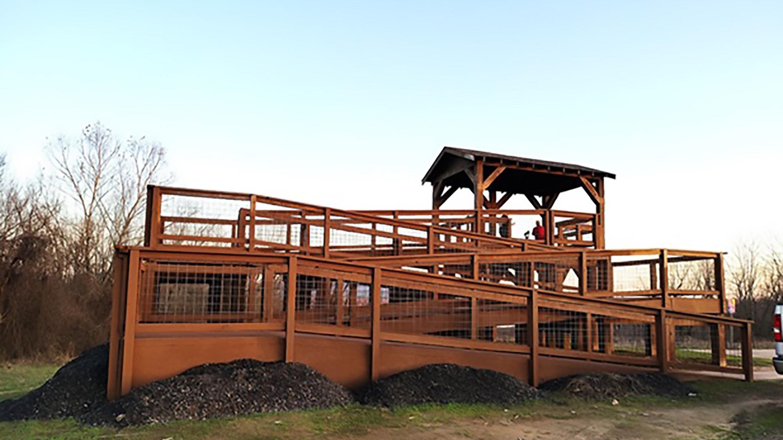 A covered wooden structure with a ramp rises above the parking lot at sunset. People stand on top.