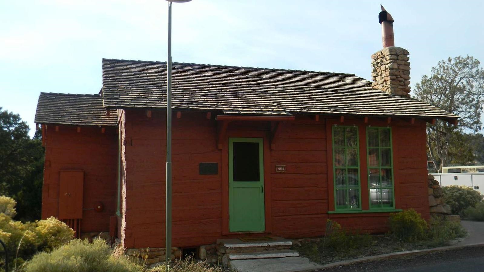A small cabin with orangish red paint, a red door, and a brown single roof with a stone chimney.