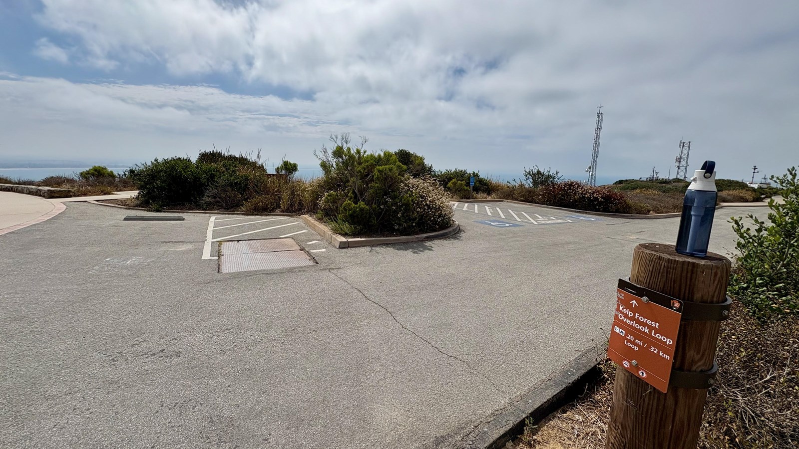 Empty parking lot, trail signpost with water bottle, bushes, towers, and ocean in the distance.
