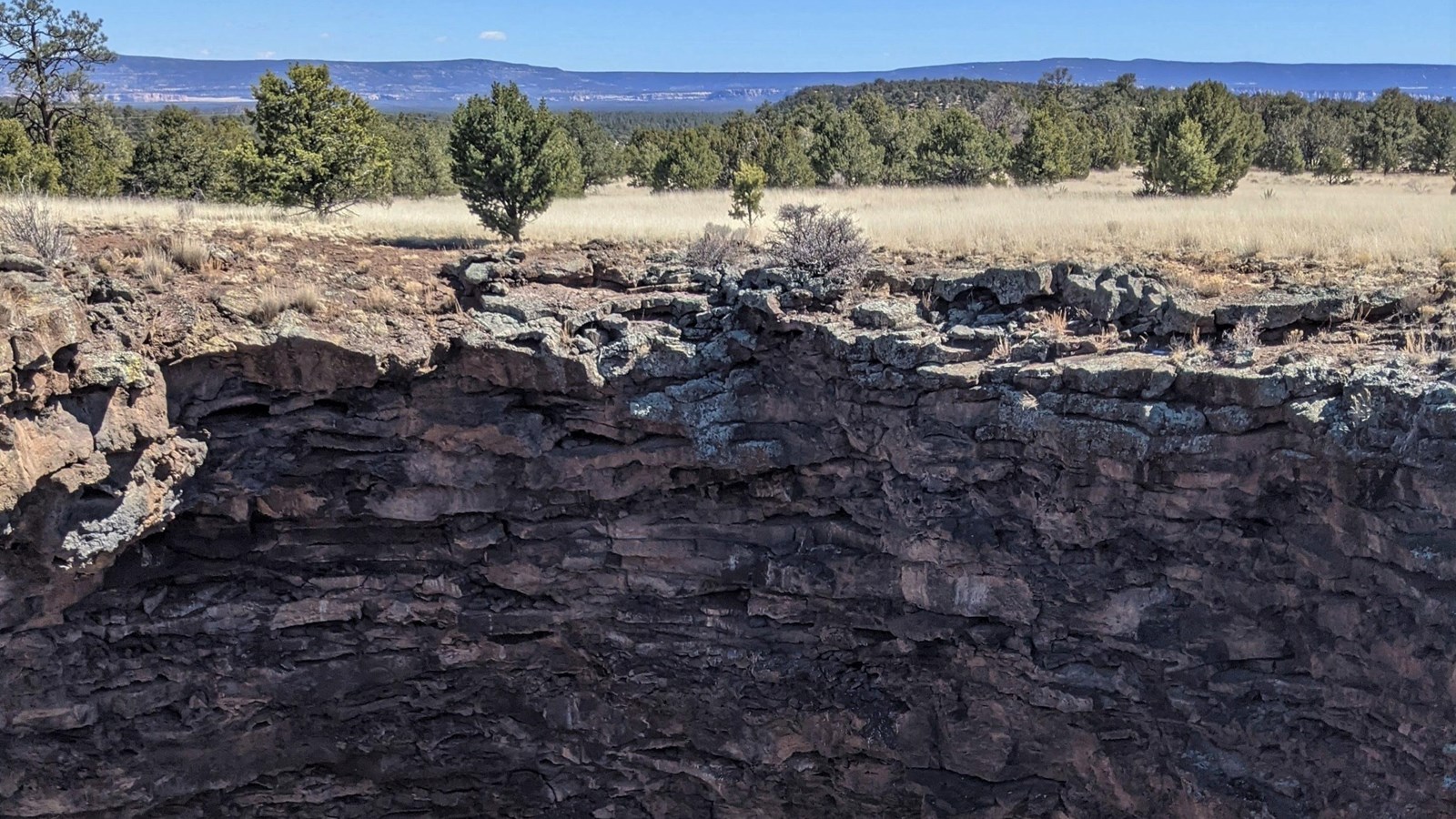 A deep pit of volcanic rock in the middle of a grassy field with shrubs and a forested ridge in the