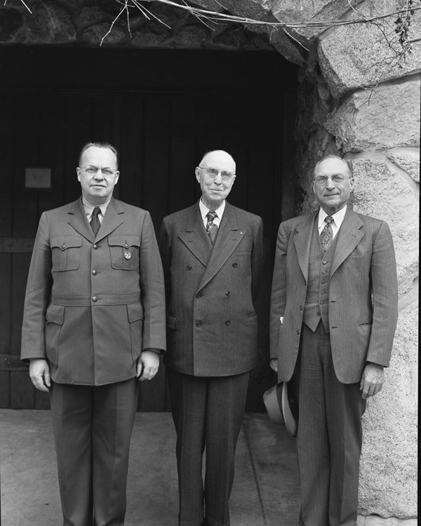 Three white men in NPS uniforms pose in front of stone building