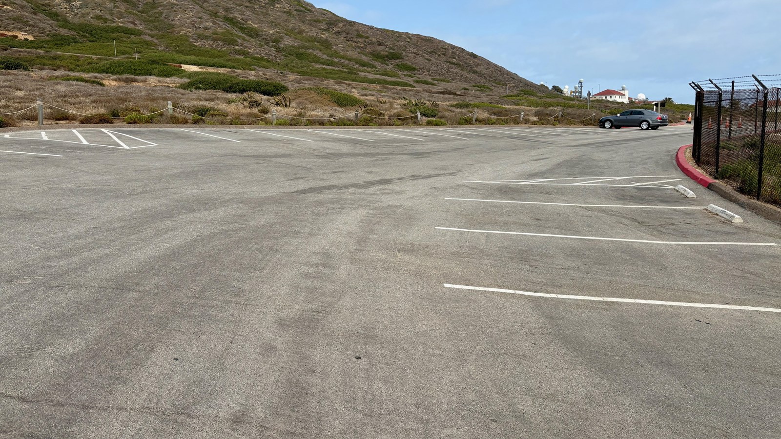 Nearly empty parking lot, one car parked, hills and a building with antennas in the background.
