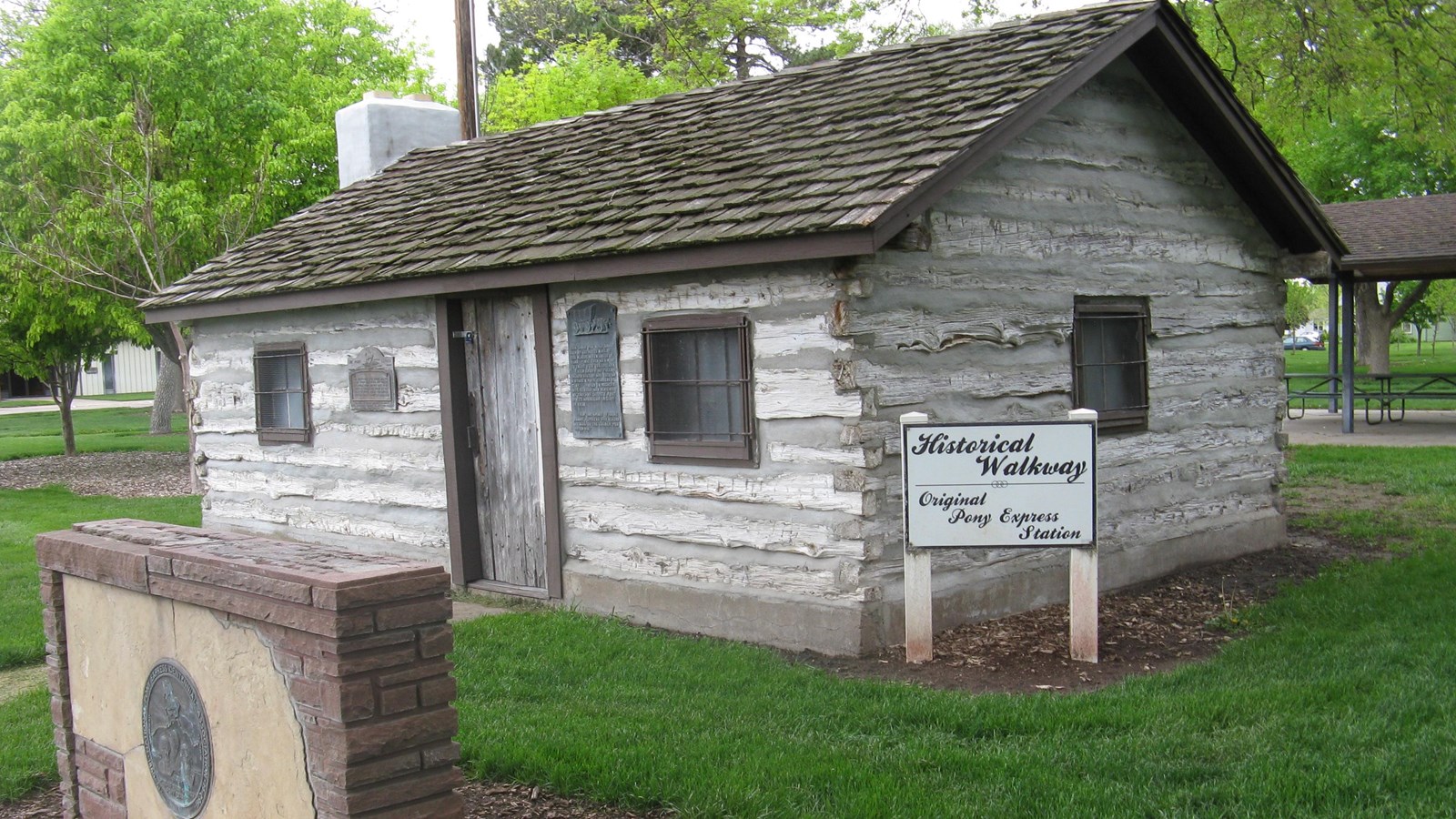 Monument and log Pony Express cabin at Cozad park, with plaques, sign, trees, picnic shelter.