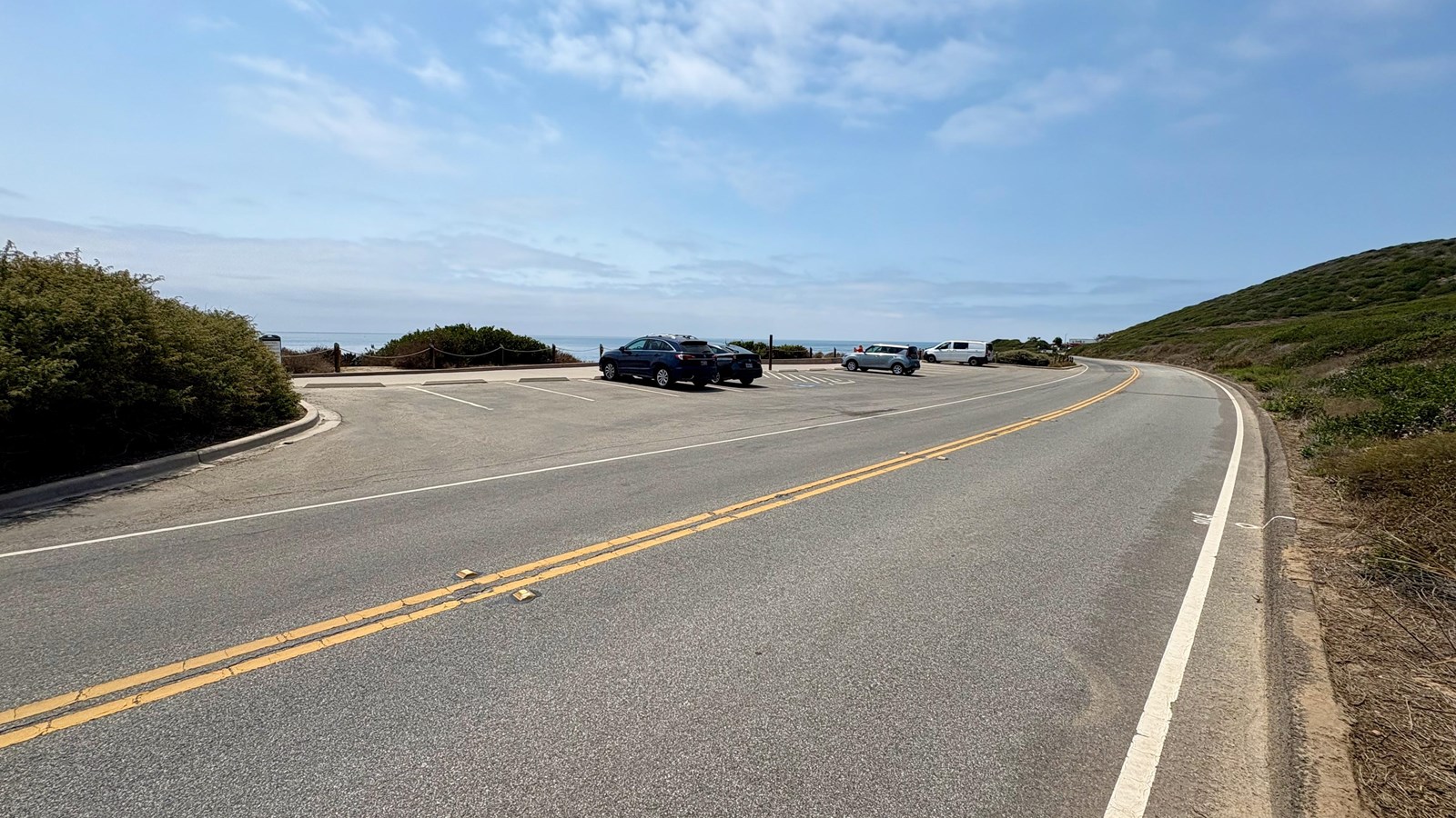 Curving coastal road, small parking lot with a few cars, ocean and blue sky in the background.