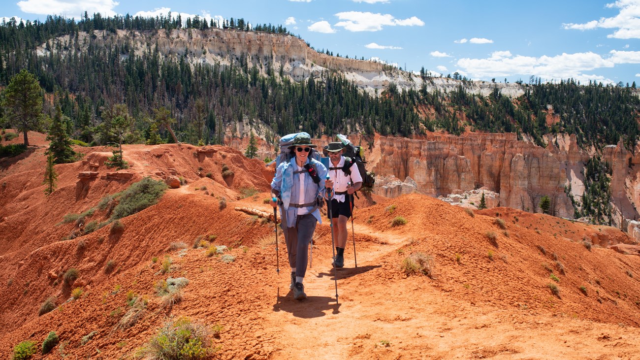 Two people walk a trail of red earth wearing gear on their backs.