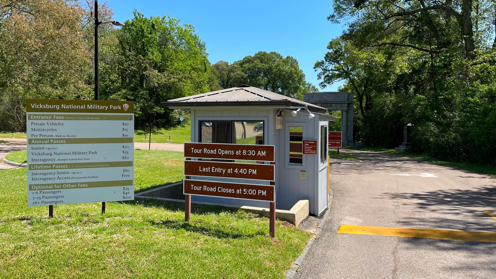 A gray small gray building with a sign showing entrance fees to the left, and a road to the right