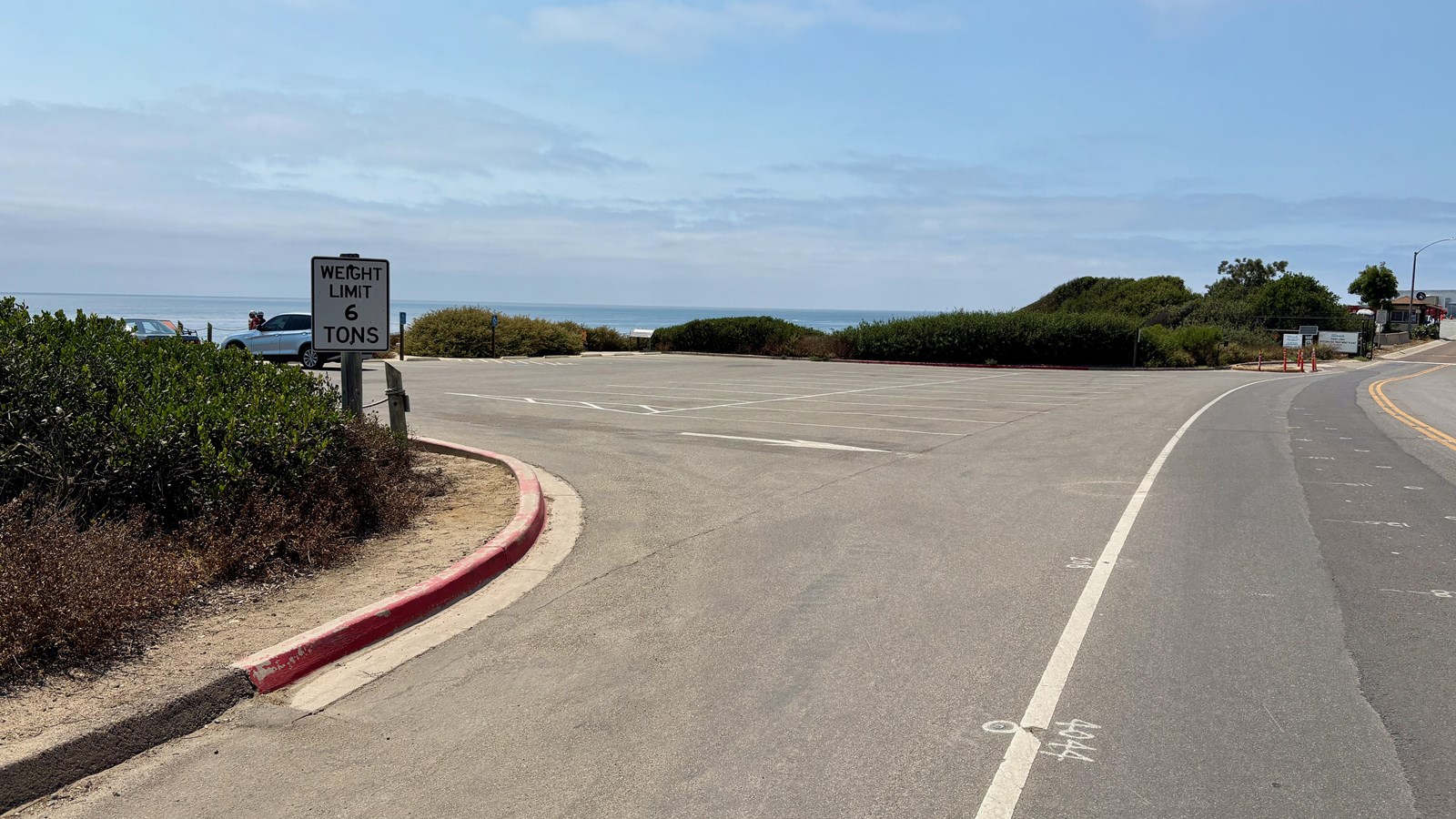 Empty parking lot by the ocean, weight limit sign, red curb, bushes, and a partly cloudy sky.