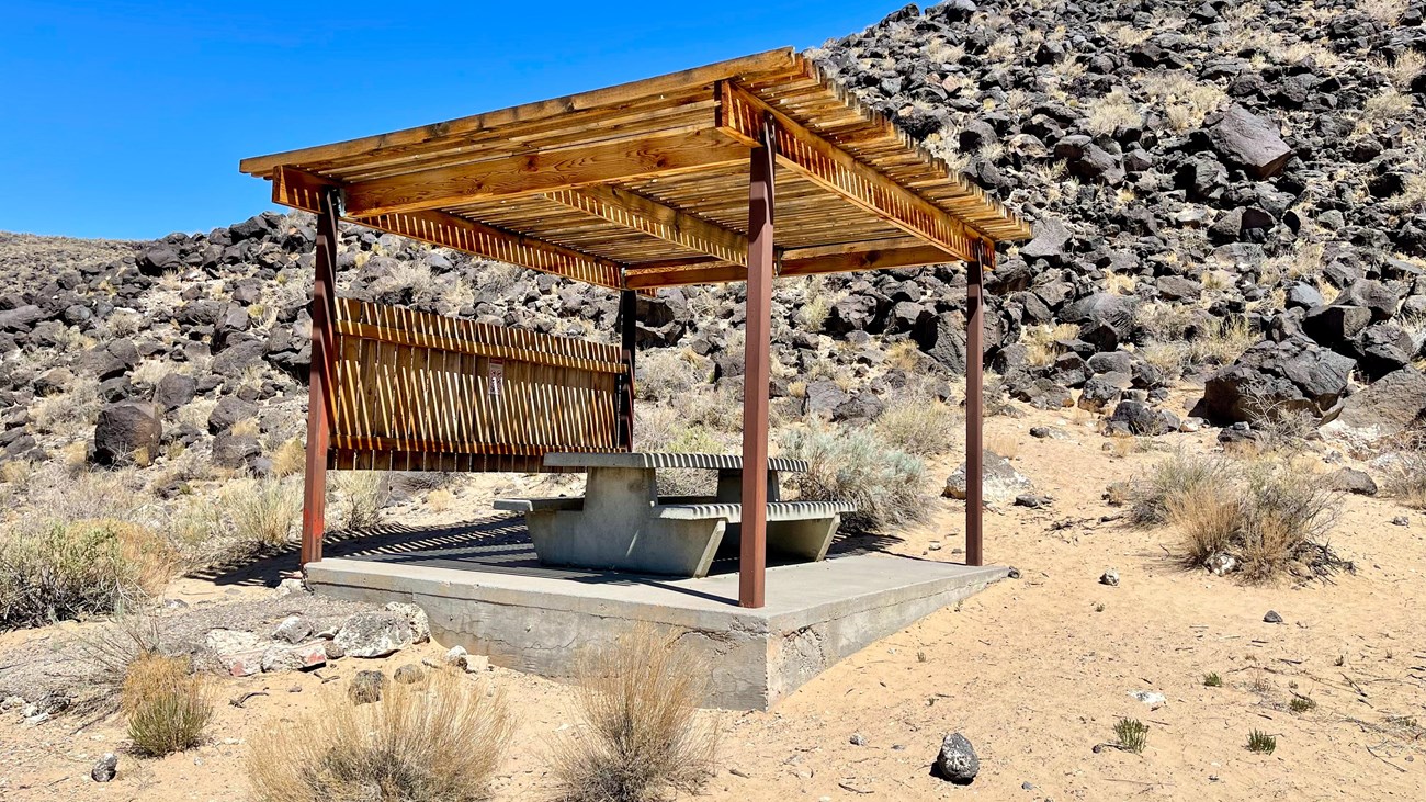 A shaded picnic pavilion with table with a lava rock escarpment in the background.