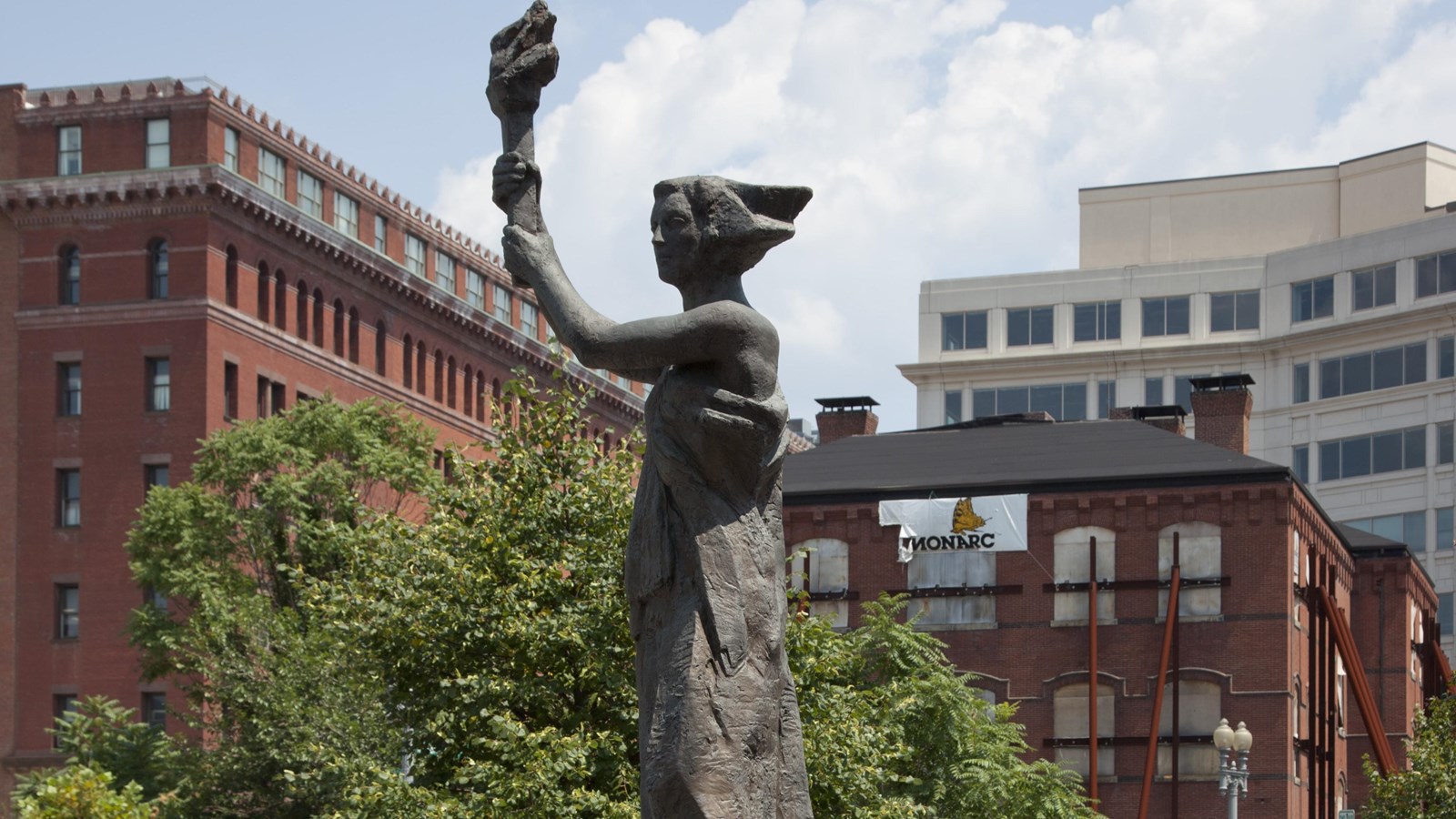 Bronze statue of a woman with a raised torch outdoors against a backdrop of trees & brick buildings