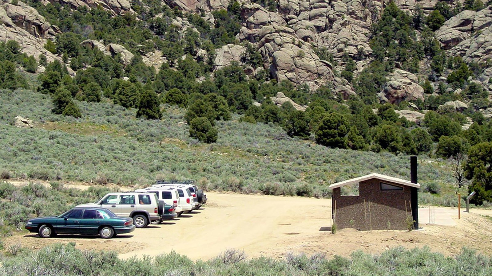 Several cars are parked in a gravel parking lot near a vault toilet near granite formations.