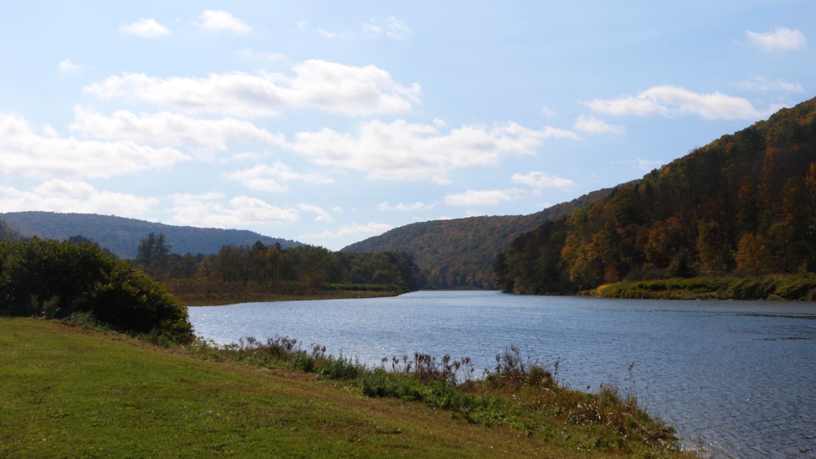 Rolling hills with fall colors around a blue river, reflecting sky. Grassy sloped bank in foreground