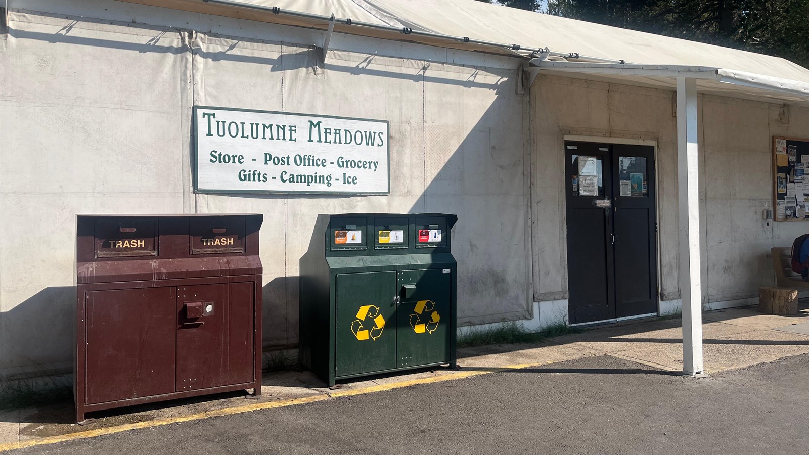 A basic white building with a tarp roof, double entrance doors, and trash and recycling outside