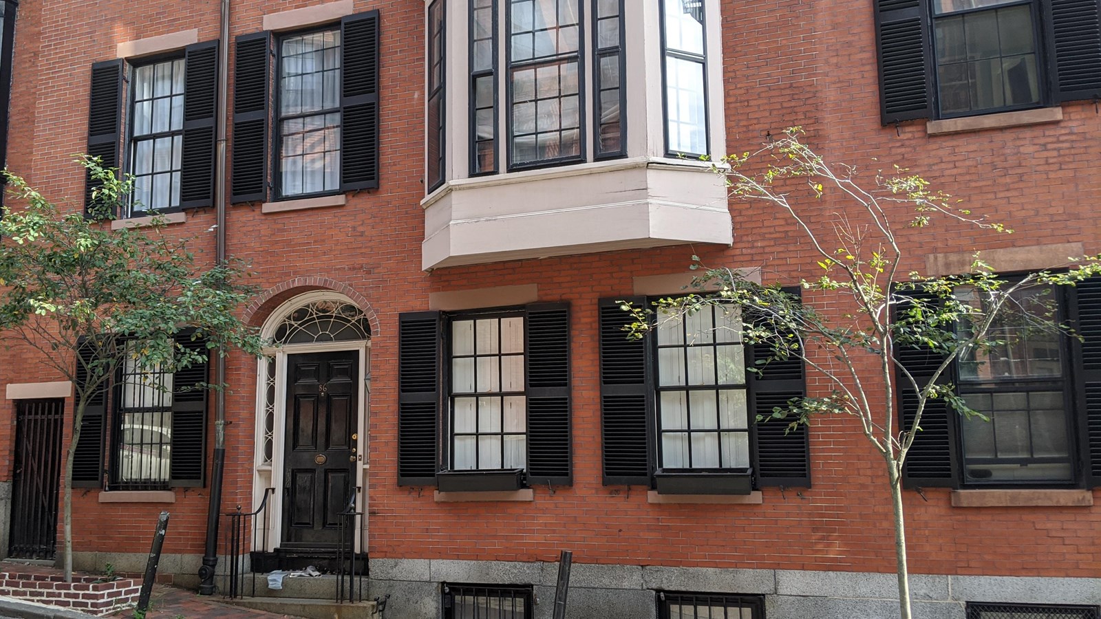 Brick building with black shutters around windows. on second floor is a white bay window.