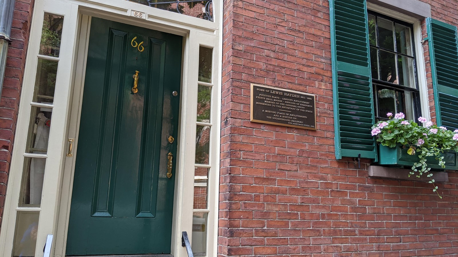 Green door and window with green shutters flank a bronze plaque on a brick wall.