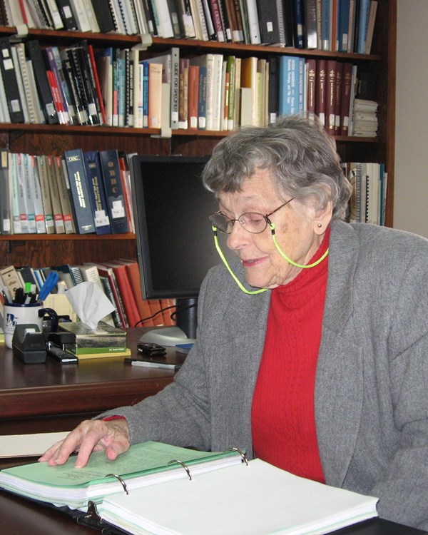 A gray-haired woman with glasses sits at a desk in a library, peering down at a binder.