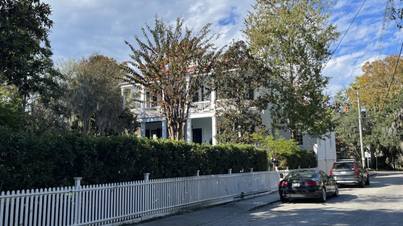 A white picket fence lines a street, alongside which a two story white house stands.