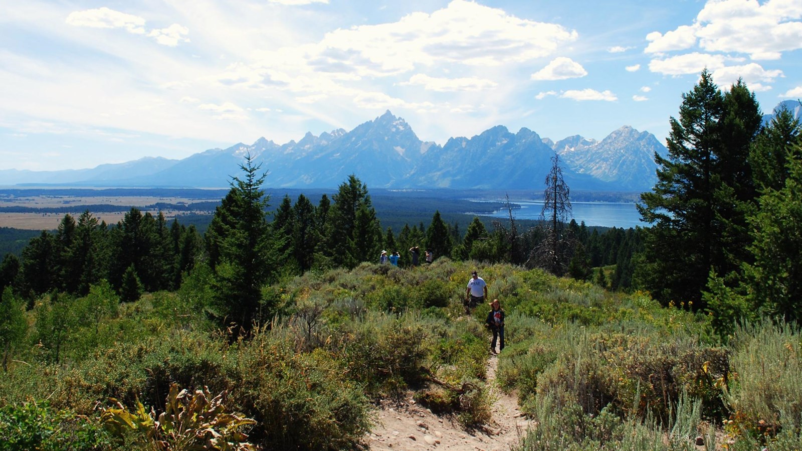 A large landscape view up high of the valley floor and Teton Range with a couple visitors.