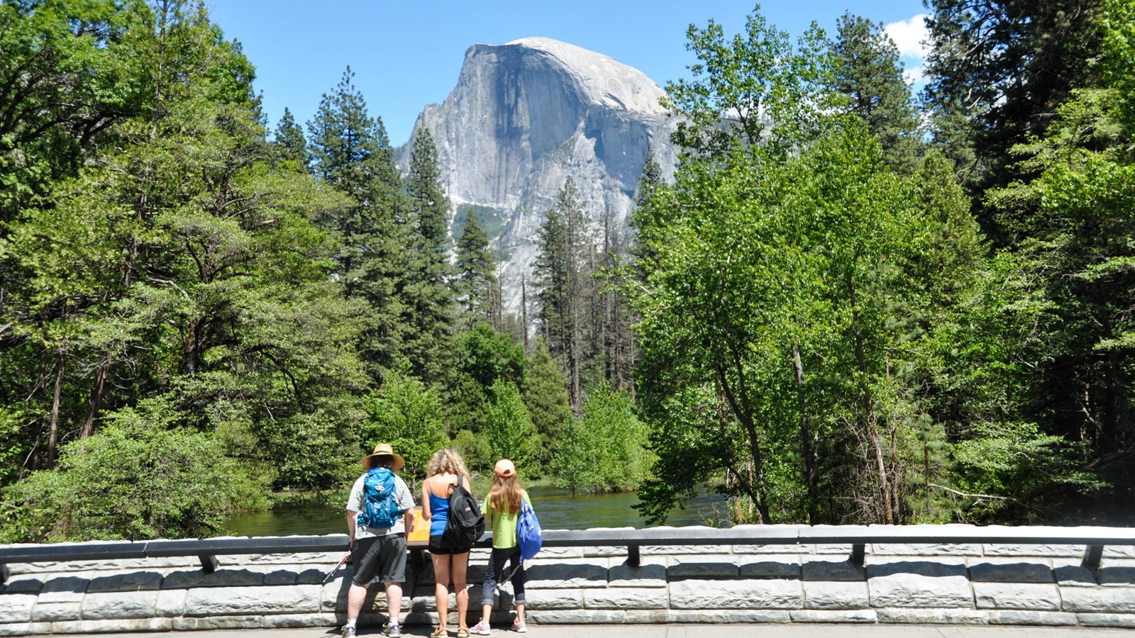 Visitors read an exhibit panel on Sentinel Bridge. Half Dome in background.
