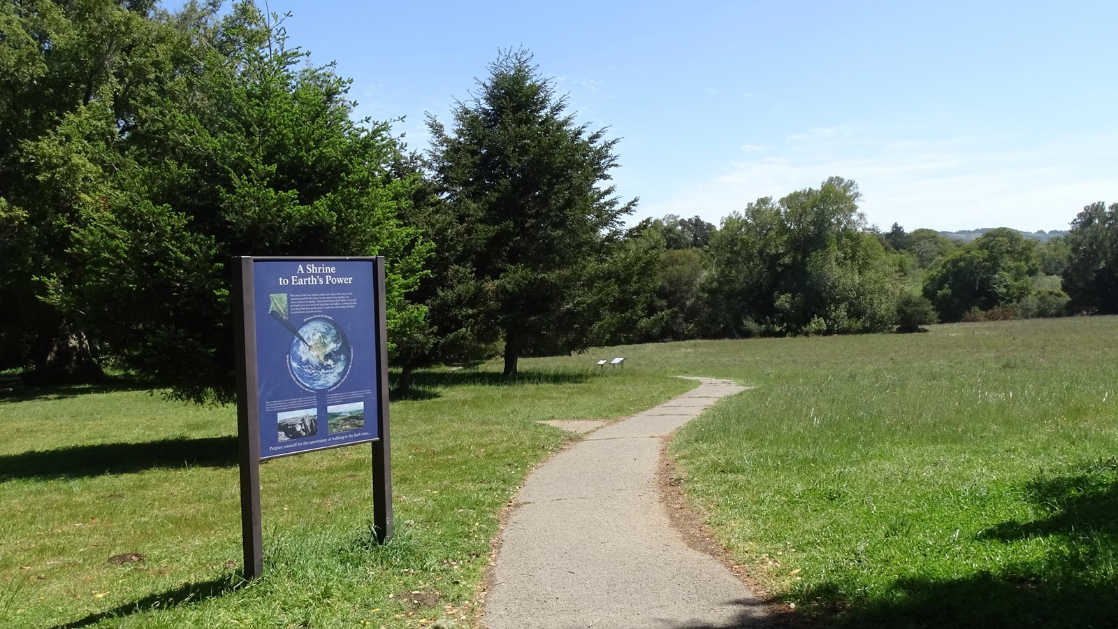 A paved trail passes to the right of a blue interpretive sign that features an image of Earth.