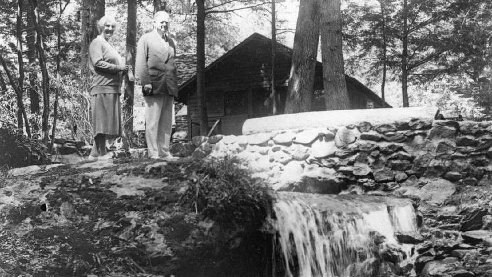 President and Mrs. Hoover stand on a large boulder to the left of a small waterfall.