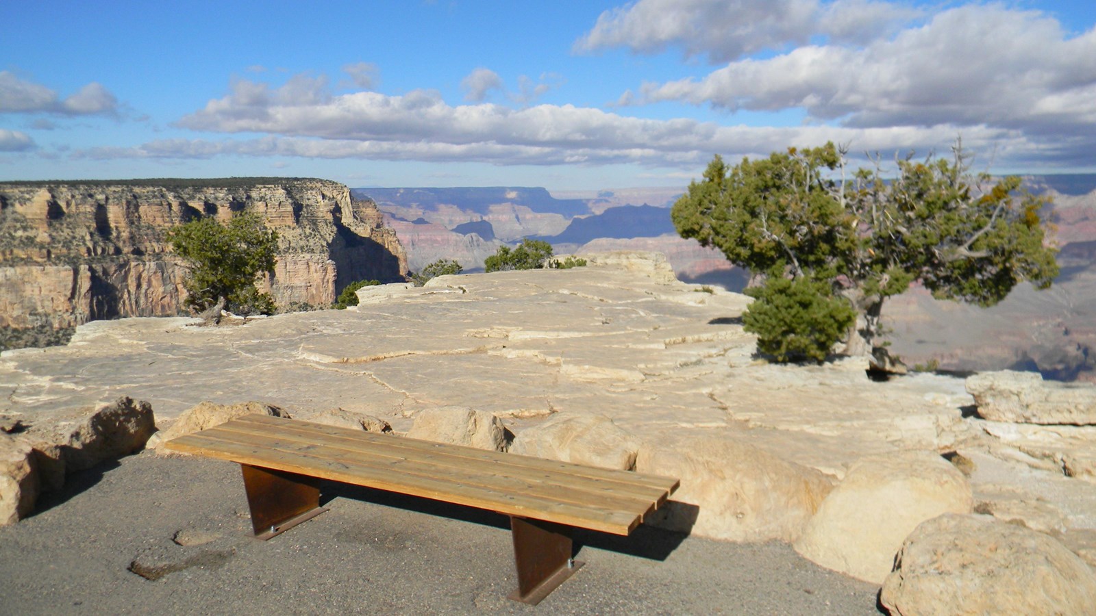 A wooden bench sits at the edge of a paved path next to a large, open outcrop of flat white rock.