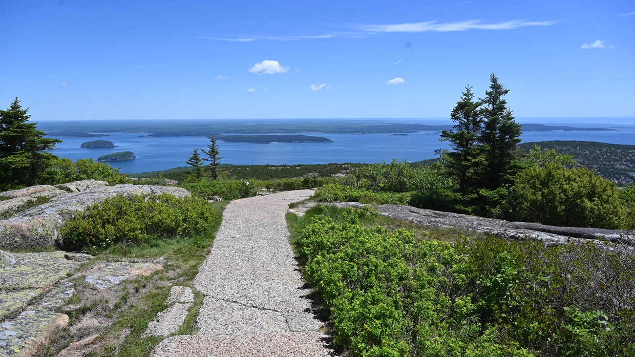 View of a paved trail on a mountain summit with islands and mountains extending to the far horizon. 