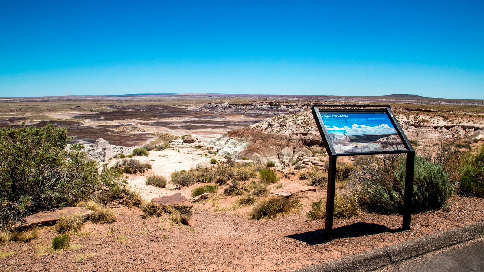 View of mesas, escarpment, floodplain, with wayside exhibit under a blue sky.