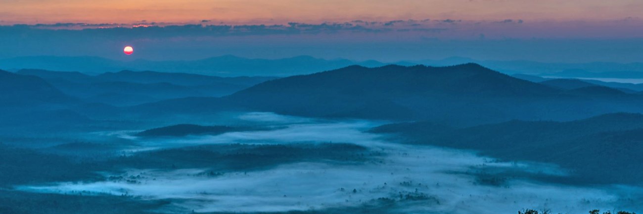 The sun rises above mist filled valleys surrounded by mountains.