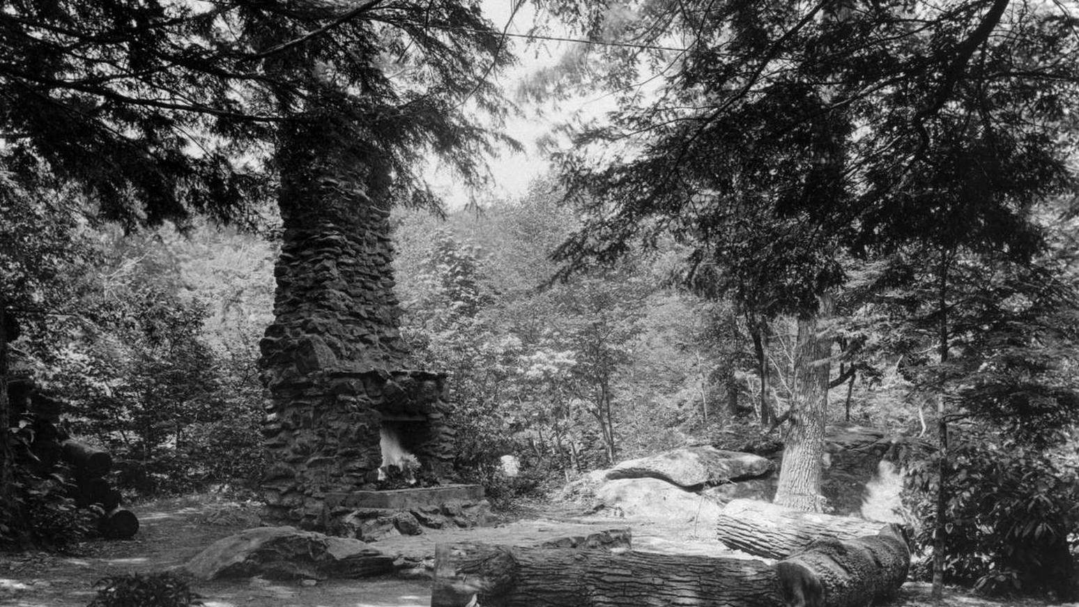 A black and white photograph of an outdoor fireplace in the woods.