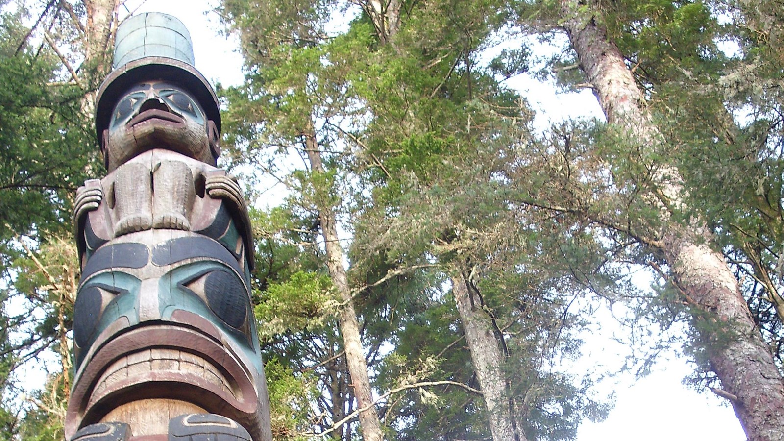 A totem pole carved in Northwest Coast style. On top is a man with a tall hat.