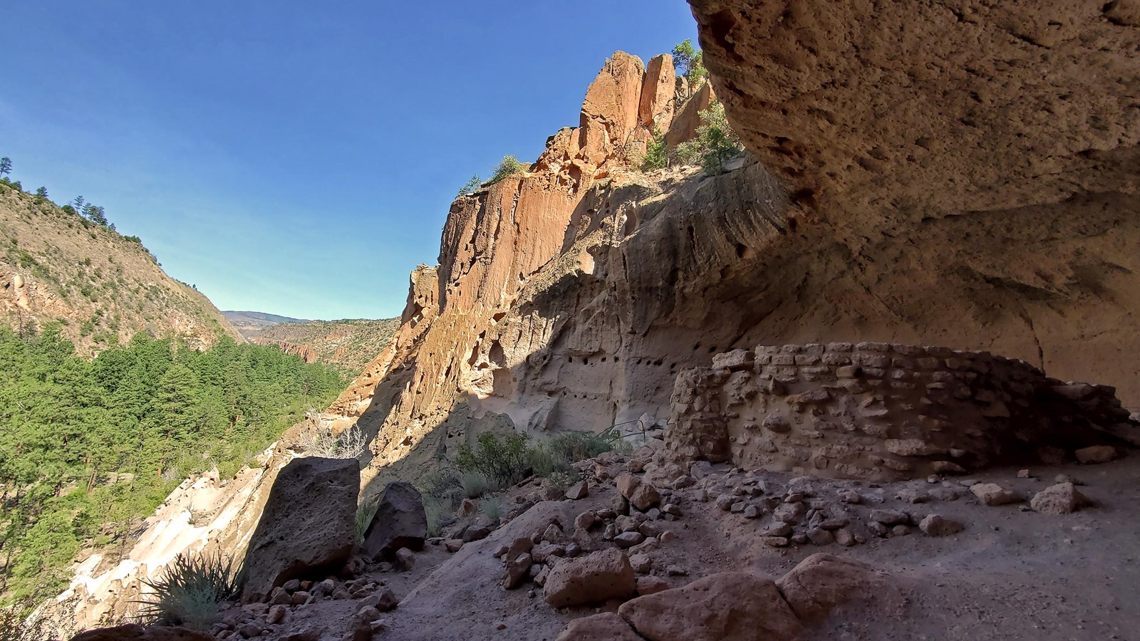 A circular stone structure sits within an alcove and a view into a green canyon with blue sky