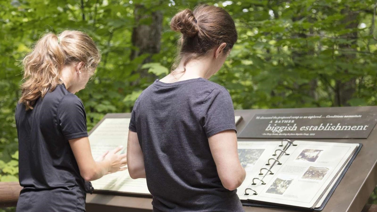 A woman and a young girl reading an outdoor exhibit with woods in the background.
