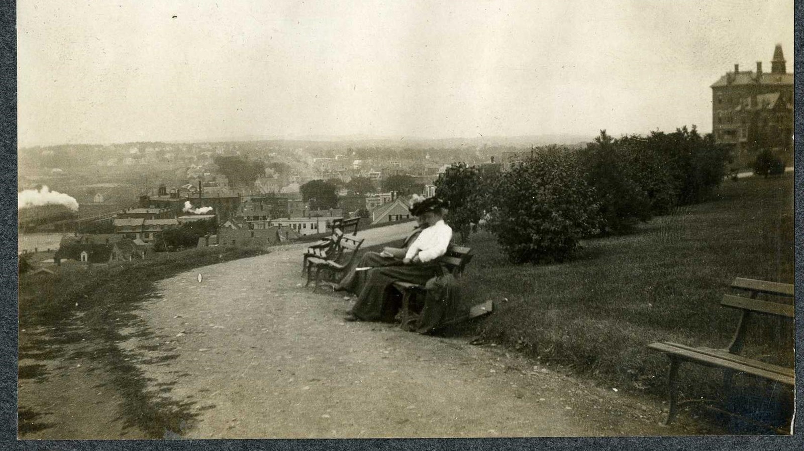 Black and white of dirt path on hill lined with benches and grass with some people sitting on bench
