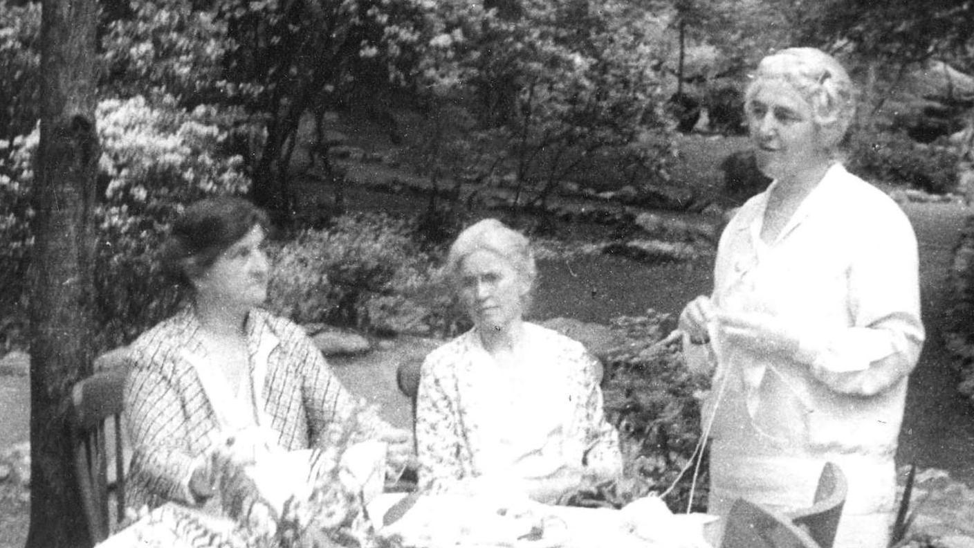 Three ladies, two seated and one standing. The lady standing is knitting.