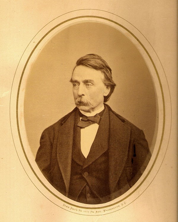 Portrait of a man with wavy short hair and a mustache, in a dark-colored three piece suit.